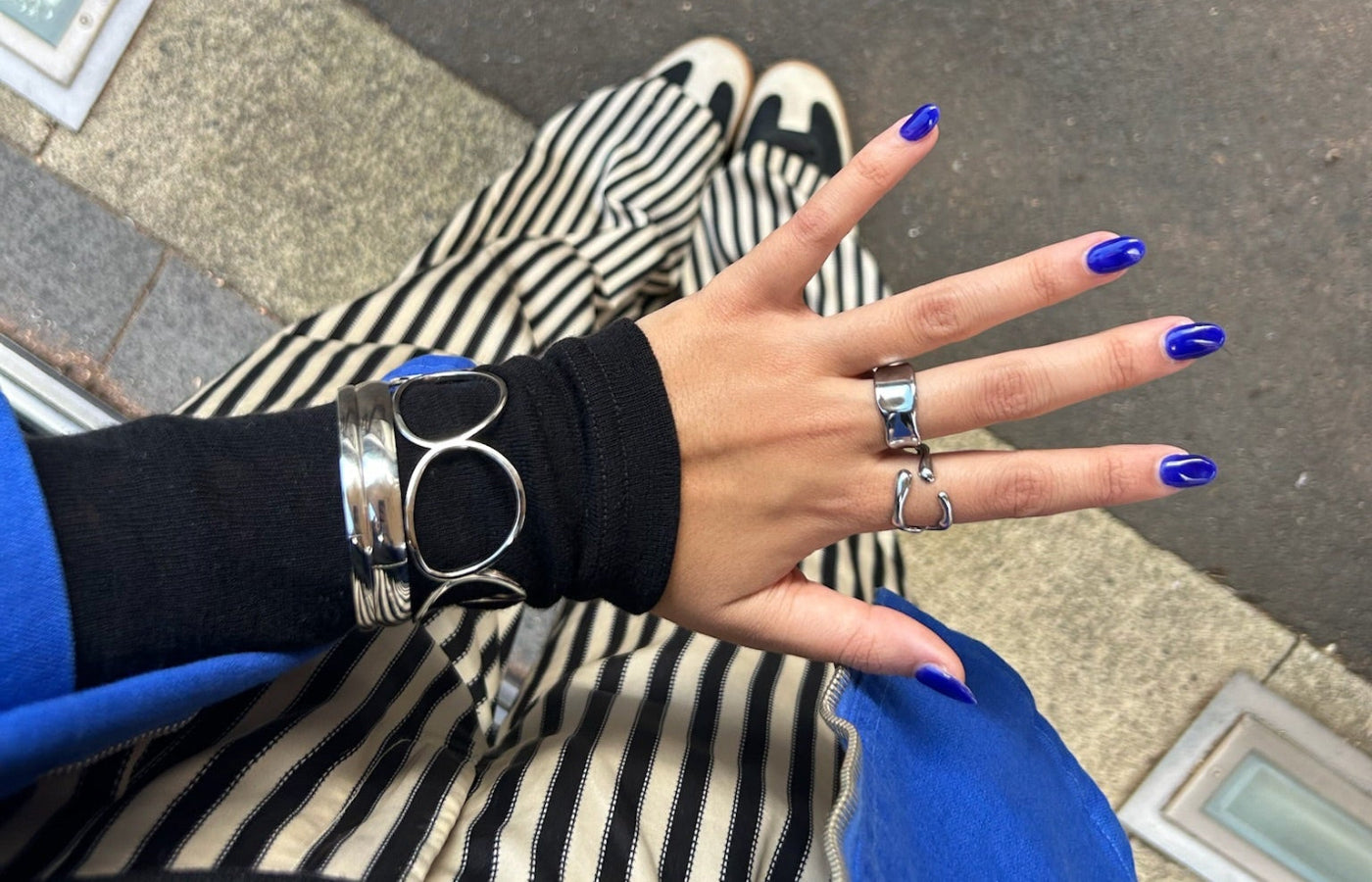 Hand with blue nail polish wearing multiple rings and a black sleeve with a silver bracelet on a striped background.