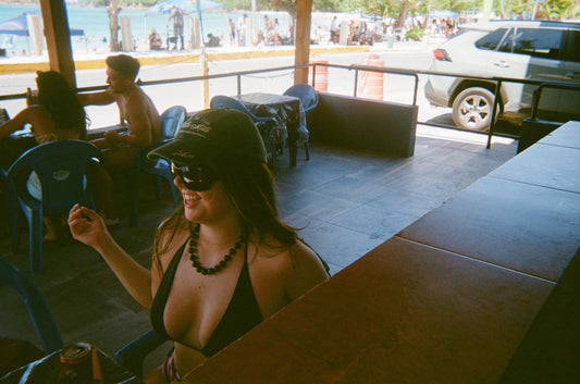 Woman in a bikini top and sunglasses sitting at a table in a casual outdoor setting wearing Mimi Necklace in Olive Green