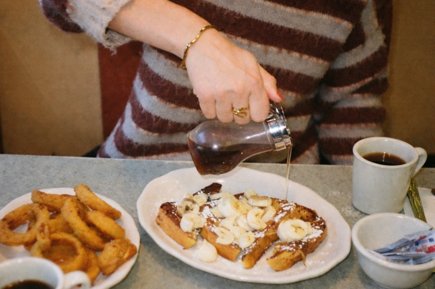 Person pouring syrup onto a plate of waffles with a side of onion rings and a cup of coffee. Roshe Ring