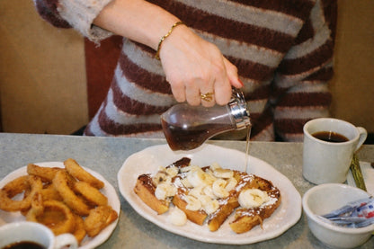 Person pouring syrup onto a plate of waffles with a side of onion rings and a cup of coffee. Roshe Ring