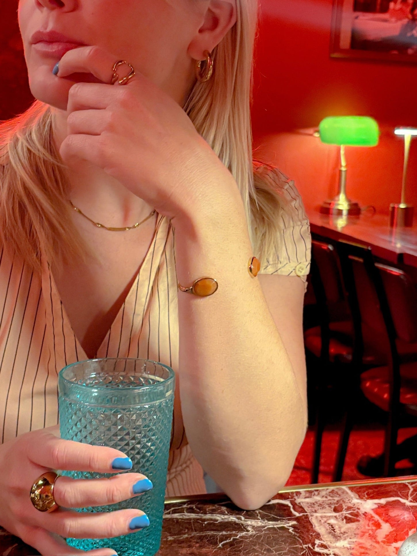 Woman holding a blue glass in a bar setting with red walls and a green lamp wearing the Sarah Ring and the Madison Cuff - Burnt Orange