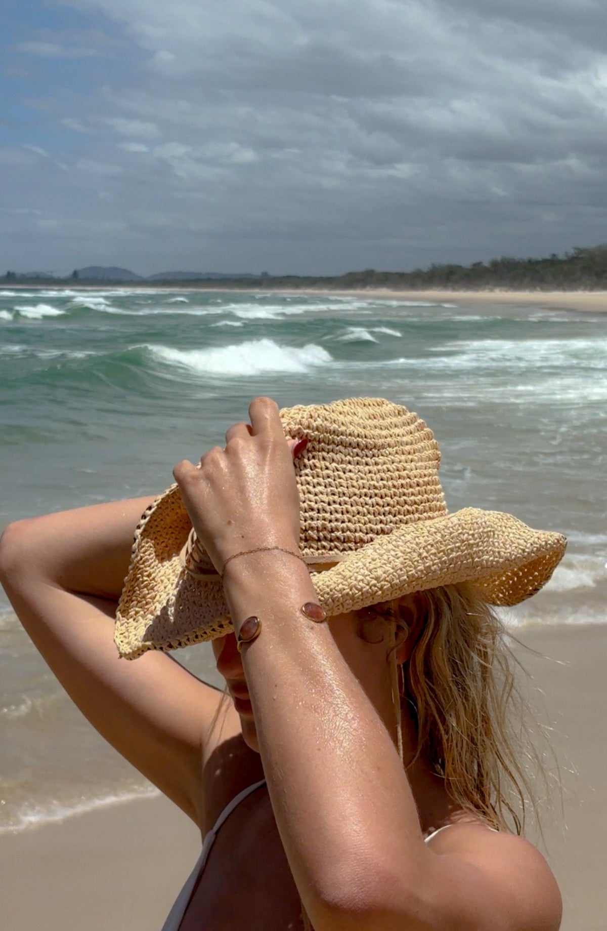 Person on a beach holding a straw hat, with ocean waves and cloudy sky in the background wearing the madison cuff